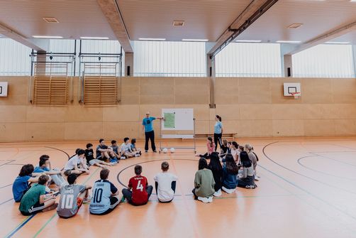Lehrer zeigt vor Schülern und Schülerinnen auf Plaktat in einer Sporthalle  
