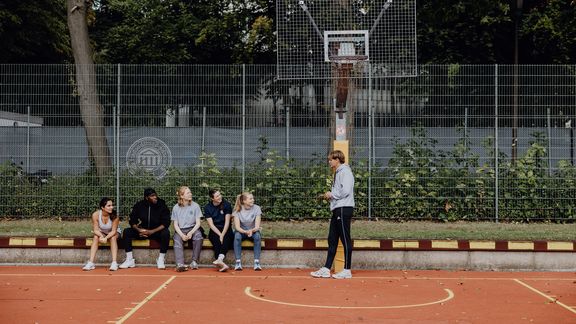 Foto von Personen die unter Basketballkorb sitzen und einer stehenden Person beim Reden zugucken
