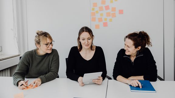 Foto von drei Frauen die am Tisch sitzen und auf ein Tablet in der Mitte blicken
