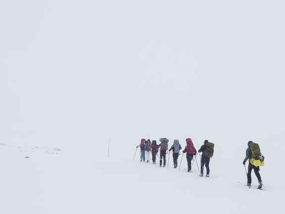 Gruppe von Schneewanderern mit Rucksäcken und Wanderstöcken in einer weißen Schneelandschaft bei eingeschränkter Sicht
