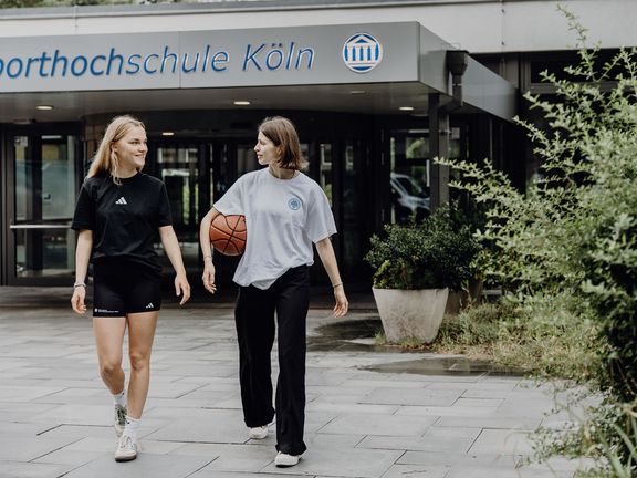 Zwei Studentinnen kommen aus Richtung des Eingangs der Deutschen Sporthochschule
