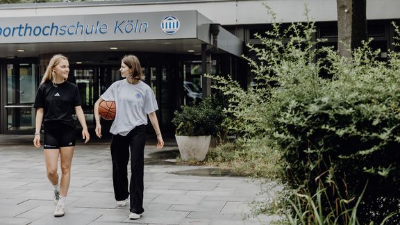 Zwei Studentinnen kommen aus Richtung des Eingangs der Deutschen Sporthochschule

