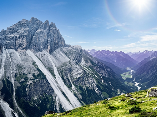 Berglandschaft mit schroffen Gipfeln, grünem Tal und Wanderer neben Zelt auf Wiese
