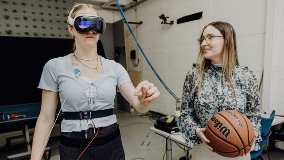Probandin mit VR-Brille zeigt in leeren Raum, während forschende Person neben ihr einen Basketball hält
