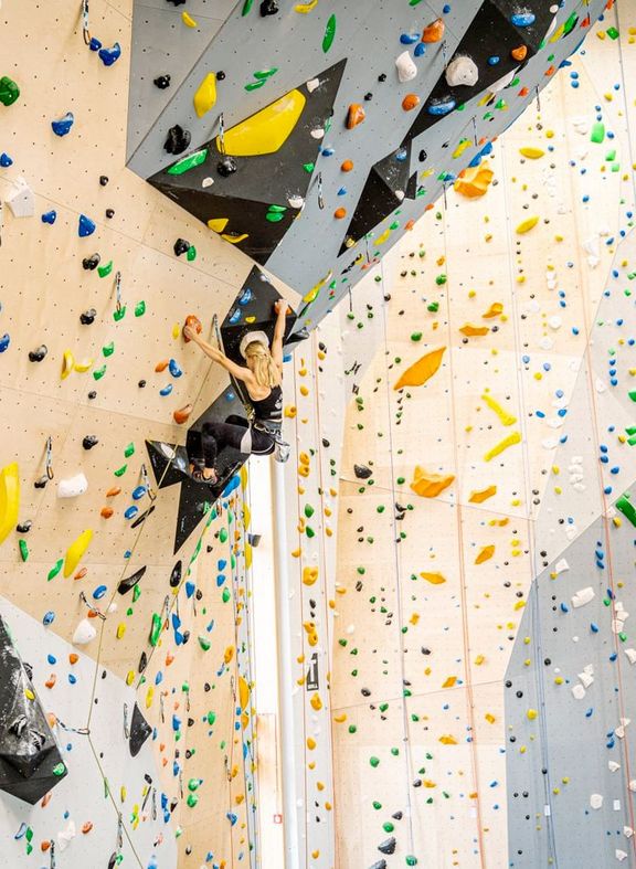 Frau klettert an einer bunten Kletterwand in einer Halle mit Vorstiegssicherung
