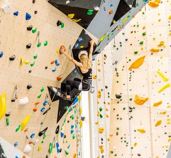 Frau klettert an einer bunten Kletterwand in einer Halle mit Vorstiegssicherung
