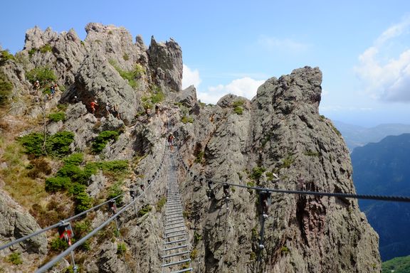 Hängebrücke aus Metallseilen und Holzplanken führt über felsiges Gebirge mit mehreren Kletterern
