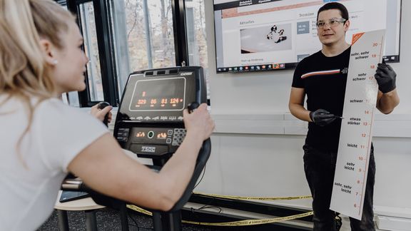 Foto von forschender Person der einer auf dem Ergometer sitzenden Probandin eine Skala zeigt
