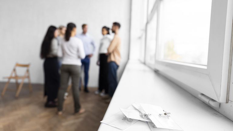 Gruppe von sechse personen leicht verschwommen im hintergrund des bildes im kreis stehend; im vordergrund weiße fensterbank, auf der namensschilder liegen
