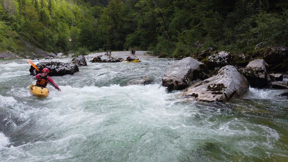 Ein Kajakfahrer mit rotem Helm und gelber Ausrüstung paddelt durch einen Wildwasserfluss, der von großen Felsen durchzogen ist. Im Hintergrund sind weitere Personen auf dem Wasser zu sehen. Dichte, grüne Bäume säumen das Ufer, und das Wasser ist klar und schäumend.
