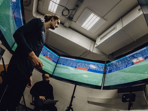 Foto von Mann der in der Mitte von sechs kreisförmig aufgestellten Bildschirmen mit Fußballsimulation steht

