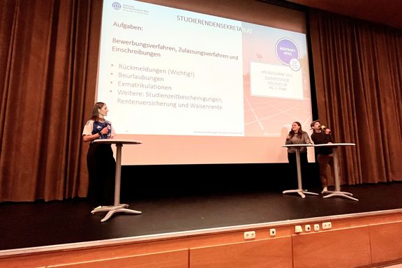 Auf der Bühne eines Hörsaals steht links eine Moderatorin und rechts stehen zwei Referent*innen. Im Hintergrund ist eine große Leinwand mit einer laufenden Präsentation zu sehen. 
