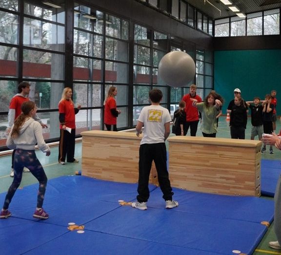 Schüler*innen werfen einen Gymnastikball über zwei Turnkästen
