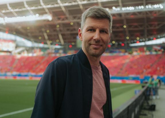 Thomas Hitzelsperger. Ein Mann mit schwarzem Jacket, lächelt in die Kamera. Im Hintergrund sind die Tribünen eines Stadions zu sehen.
