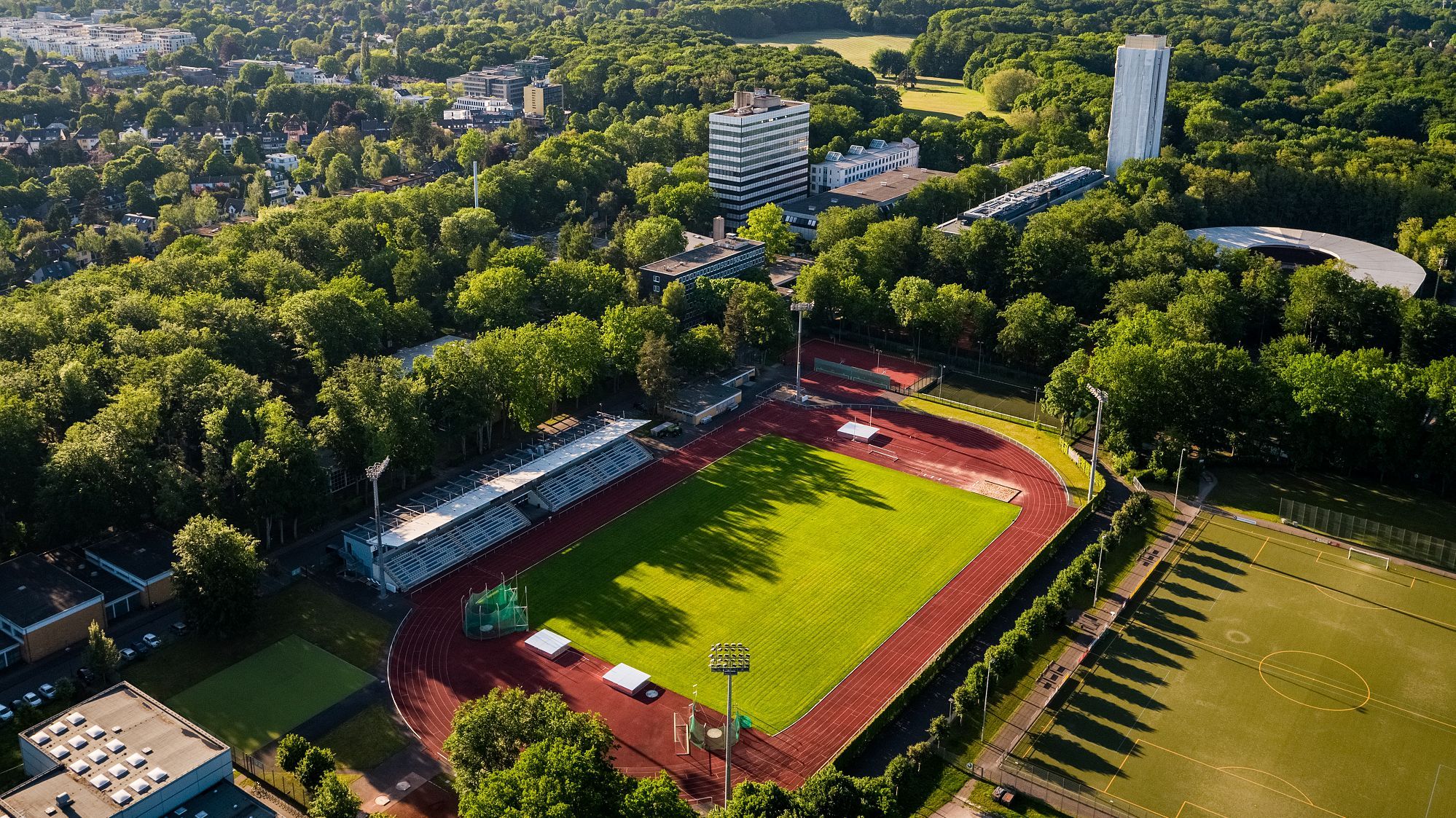 Eine Luftbildaufnahme eines Sportplatzes mit umlaufender Aschenbahn umringt von vielen Bäumen.
