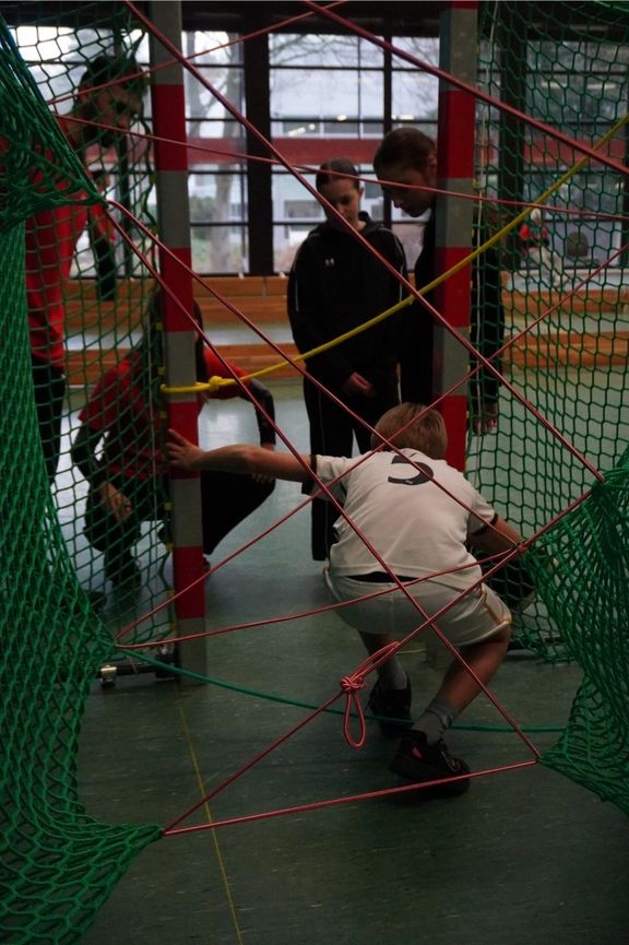 Mehrere Kinder bewegen sich in einer Sporthalle durch ein Parcours-Element aus gespannten roten Seilen und grünen Netzen. Ein Kind kriecht geduckt unter den Seilen hindurch, während andere Kinder daneben stehen und den Parcours beobachten
