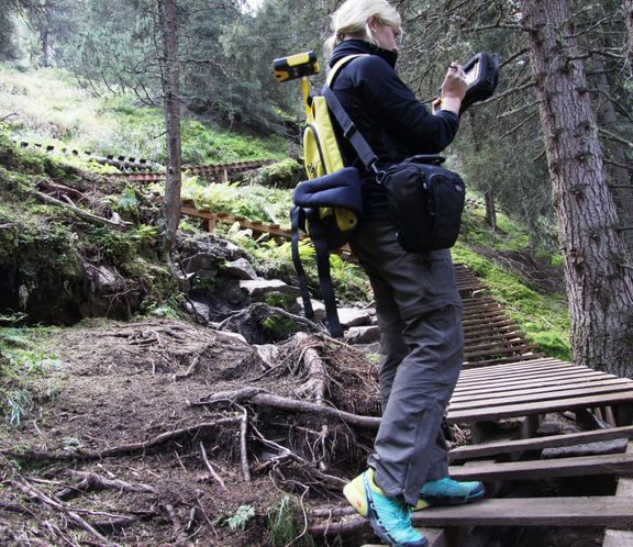 Eine blonde Frau steht auf einem Wanderweg aus Holz im Wald und tippt auf ein Forschungsgerät. Auf dem Rücken trägt sie ebenfalls Forschungsequipment. Sie trägt Wanderkleidung.
