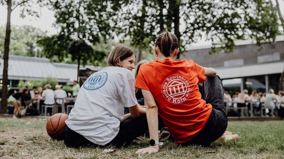 Studentinnen sitzen mit dem Rücken zur Kamera auf dem Gras und reden miteinander
