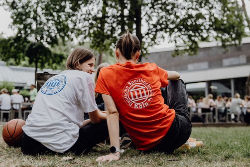 Studentinnen sitzen mit dem Rücken zur Kamera auf dem Gras und reden miteinander
