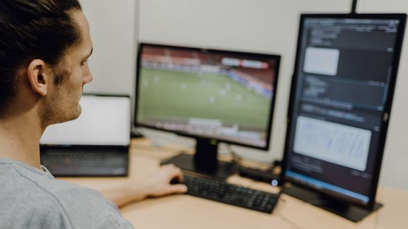 Foto von Mann der auf auf Programmiercode neben laufendem Fußballspiel blickt
