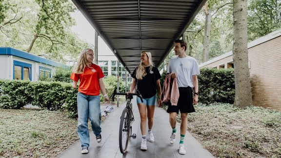 Drei Studierende gehen mit einem Fahrrad durch die Gänge am Campus
