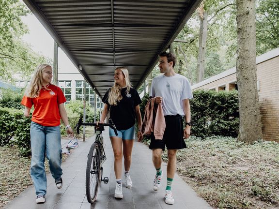 Drei Studierende gehen mit einem Fahrrad durch die Gänge am Campus
