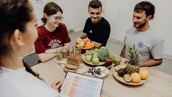Frau erklärt Gruppe Ernährungstabelle, Auf dem Tisch steht Obst und Gemüse
