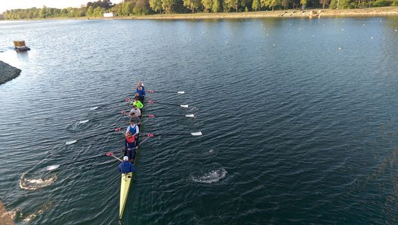 Ein Ruderboot mit mehreren Personen bewegt sich auf einem ruhigen Gewässer. Die Ruderblätter sind gleichmäßig ins Wasser getaucht, wodurch kreisförmige Wellen entstehen. Am Ufer im Hintergrund stehen zahlreiche Bäume, das Wasser wirkt tiefblau. Die Szene ist aus erhöhter Perspektive aufgenommen.
