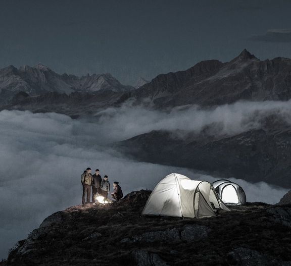 Eine Gruppe Menschen steht auf einem Berggipfel um ein Licht herum, vor ihnen steht ein erleuchtetes Zelt.
