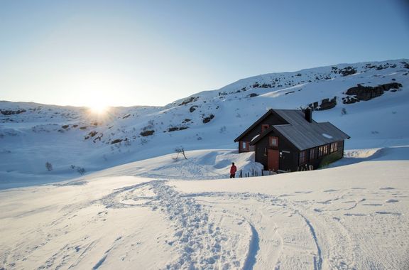 Einsames Haus in verschneiter Berglandschaft bei Sonnenaufgang mit einer Person davor
