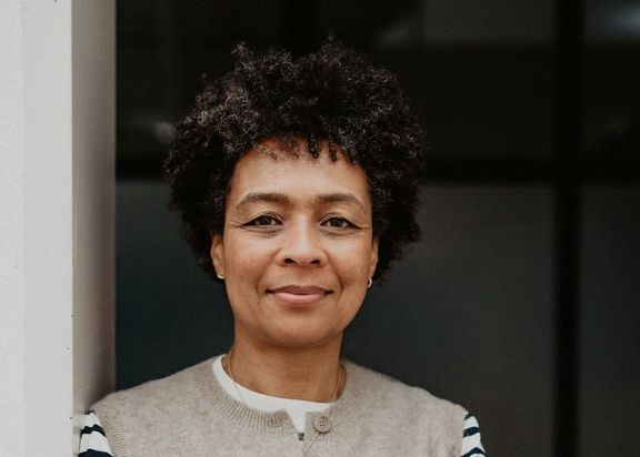 Porträtfoto von Susanne Amar. Sie steht an eine weiße Wand angelehnt vor einem dunklen Hintergrund. Sie trägt ein weiß-schwarz gestreiftes Langarm-Oberteil mit einer grauen ärmellosen Jacke darüber.
