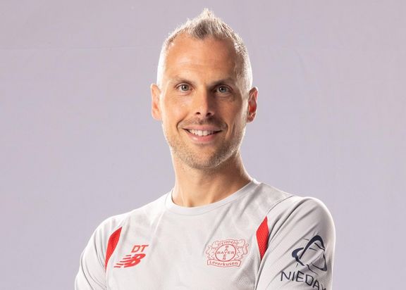 Porträtfoto von David Thiel. Er trägt ein weißes Bayer 04 Leverkusen-Trikot und steht vor einer grauen Wand.
