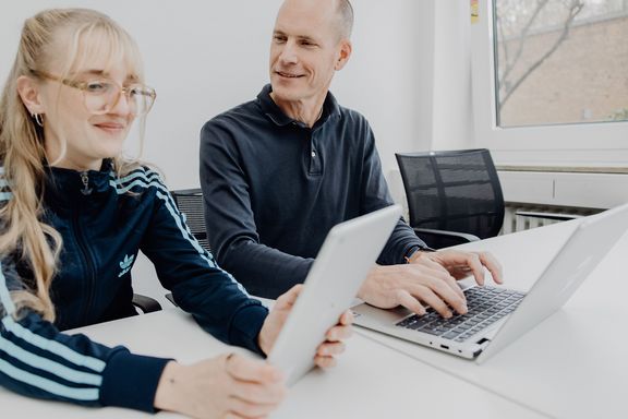 Foto von zwei Personen die an einem Tisch sitzen und mit Laptop und Tablet arbeiten
