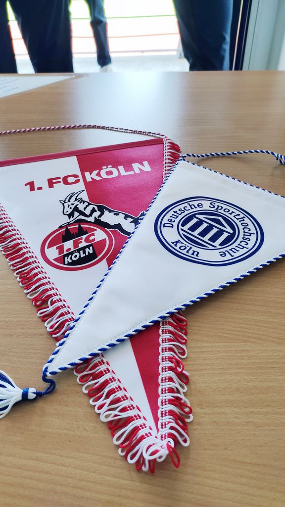 2 Wimpel liegen schräg übereinander, der untere größere zeigt das Emblem des 1. FC Köln in Rot und Weiß und der obere kleinere Wimpel zeigt das DSHS-Siegel in Blau auf Weiß

