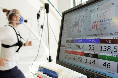Ein Sportler läuft auf einem Laufband während eines Spiroergometrietests. Er trägt eine Atemmaske und Sensoren, die seine Leistungsdaten messen. Im Vordergrund zeigt ein Monitor Diagramme, Kurven und Messwerte wie Geschwindigkeit, Herzfrequenz und Sauerstoffaufnahme in Echtzeit.
