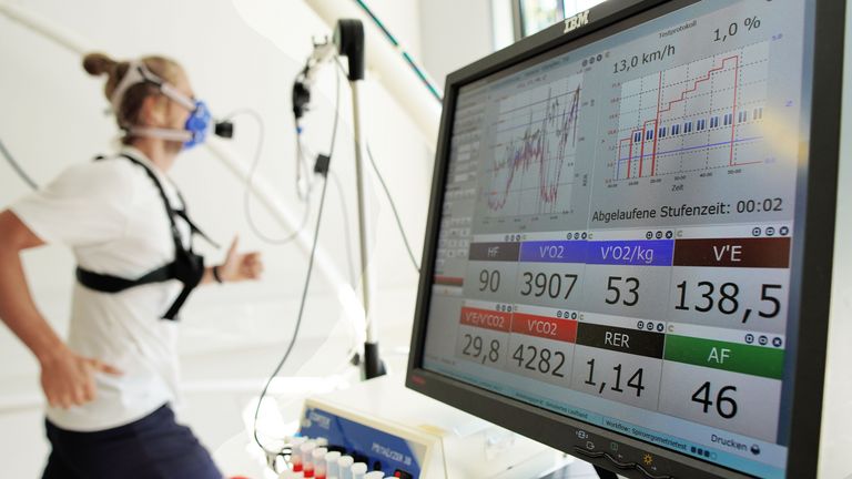 Ein Sportler läuft auf einem Laufband während eines Spiroergometrietests. Er trägt eine Atemmaske und Sensoren, die seine Leistungsdaten messen. Im Vordergrund zeigt ein Monitor Diagramme, Kurven und Messwerte wie Geschwindigkeit, Herzfrequenz und Sauerstoffaufnahme in Echtzeit.
