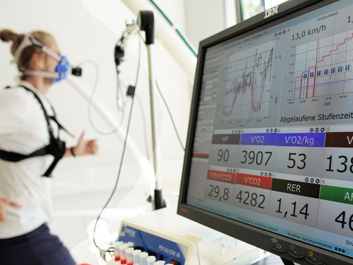 Ein Sportler läuft auf einem Laufband während eines Spiroergometrietests. Er trägt eine Atemmaske und Sensoren, die seine Leistungsdaten messen. Im Vordergrund zeigt ein Monitor Diagramme, Kurven und Messwerte wie Geschwindigkeit, Herzfrequenz und Sauerstoffaufnahme in Echtzeit.
