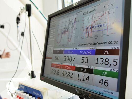 Im Vordergrund ist ein Computerbildschirm mit einer Spiroergometrie Auswertung zu sehen, während im Hintergrund eine Frau diese läuft
