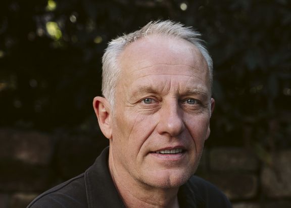 Das Foto zeigt Christian Streich, welcher vor einer Stein-Mauer sitzt und in die Kamera schaut. Er trägt ein schwarzes Hemd.
