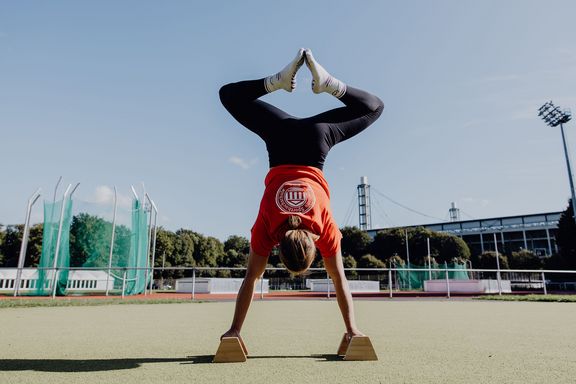 Foto einer Frau die auf zwei Holzgriffen auf dem Boden einen Handstand macht und ihre Knie stark anwinkelt
