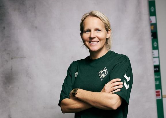 Das Foto zeigt Frederike Kromp. Eine blonde Frau mit grünem Werder Bremen T-Shirt. Sie verschränkt die Arme vor einer grauen Leinwand.
