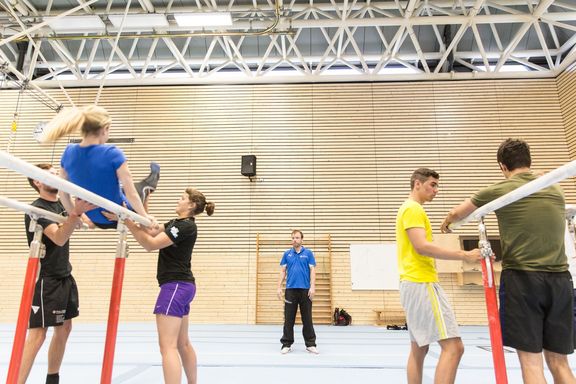 Lehrsituation in der Turnhalle, zwei Barrengeräte, links eine turnende Studentin und zwei Studentinnen, die Hilfestelllung leisten. Am rechten Barren sieht man zwei Studenten. In der Mitte der Halle steht der Dozent,.
