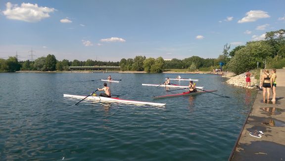 Mehrere Menschen rudern an einem sonnigen Tag mit schmalen Ruderbooten auf einem ruhigen See. Am rechten Ufer stehen einige Personen auf einem Steg und beobachten das Geschehen. Im Hintergrund sind Bäume, eine kleine Brücke und ein blauer Himmel mit vereinzelten Wolken zu sehen.
