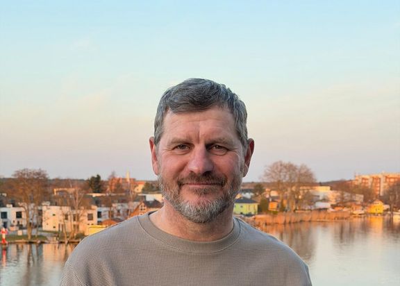 Steffen Baumgart, ein Mann mit einem beigen T-Shirt. Im Hintergrund sind ein Fluss und eine Häuserreihe am gegenüberliegenden Ufer.
