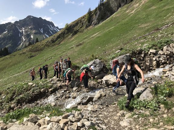 Gruppe von Wanderern überquert einen Bachlauf auf einem Bergpfad mit steinigem Untergrund und grüner Wiese im Hintergrund
