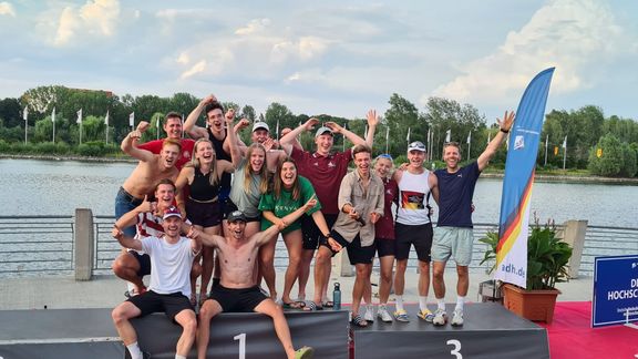 Eine große Gruppe junger Erwachsener jubelt und posiert gemeinsam auf einem Siegerpodest an einem See. Einige heben die Arme in die Luft, lachen oder zeigen Daumen hoch. Im Hintergrund sind Wasser, Bäume und Fahnen zu sehen, daneben steht eine blau-weiße Flagge einer Hochschulsportveranstaltung.
