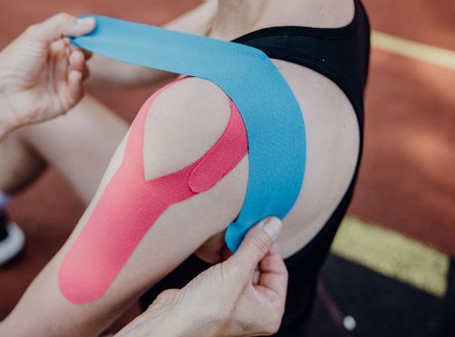 Eine Person bringt blaues Kinesiologie-Tape an der bereits mit pinkem Tape stabilisierten Schulter einer Sportlerin an.
