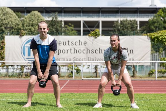 Eine Studentin und ein Student auf dem Sportplatz in leichter Hockstellung, bei halten ein Kettlebell.
