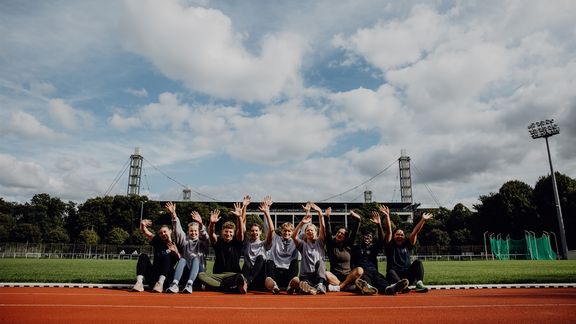 Studierende sitzen gemeinsam auf der Laufbahn des Leichtathletik-Stadions der Spoho und jubeln.
