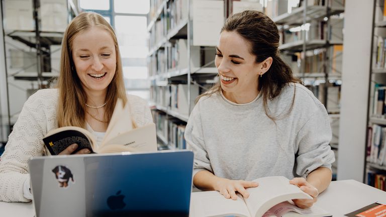 Zwei Studentinnen blättern durch ein Buch in der Bibliothek der Deutschen Sporthochschule
